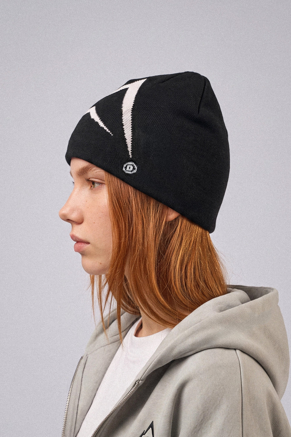 A person in profile wearing a black knit beanie with a white embroidered logo and a gray zip-up hoodie against a plain light gray background.