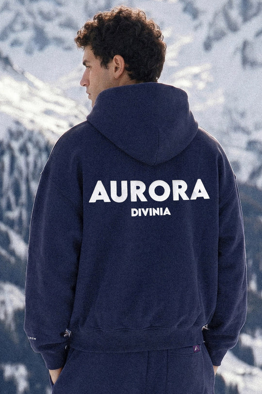 Person wearing a navy hoodie with 'Aurora Divinia' text, standing against a snowy mountain background