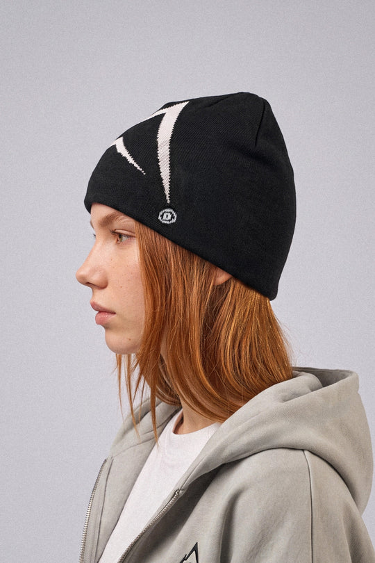 A person in profile wearing a black knit beanie with a white embroidered logo and a gray zip-up hoodie against a plain light gray background.