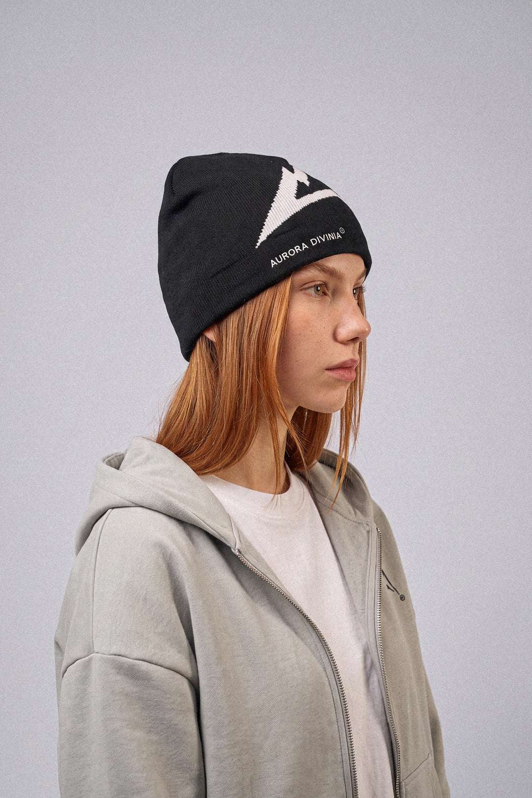 A person in profile wearing a black knit beanie with a white Aurora Borealis logo and text, paired with a light gray zip-up hoodie over a white shirt, against a plain gray studio background.