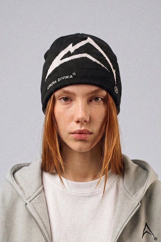 A model with orange hair wearing a black knit beanie with a white mountain graphic and the text 'AURORA DIVINIA' on the side, paired with a light gray zip-up hoodie against a solid gray background.