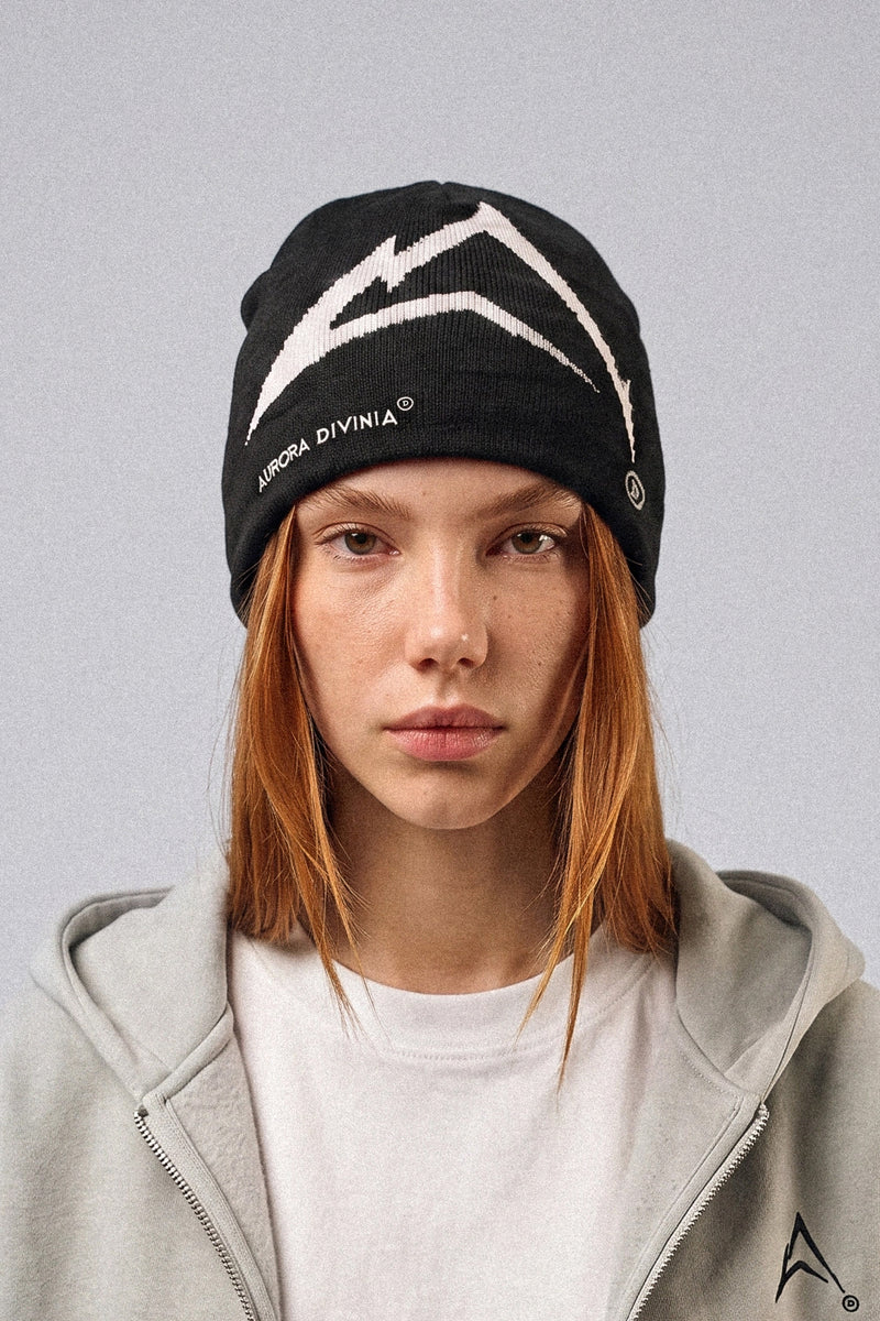 A model with orange hair wearing a black knit beanie with a white mountain graphic and the text 'AURORA DIVINIA' on the side, paired with a light gray zip-up hoodie against a solid gray background.