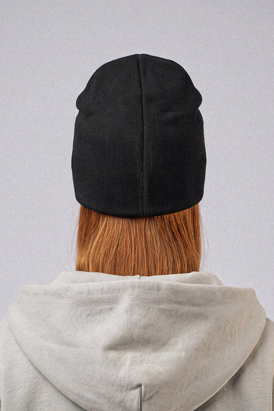 A person viewed from behind wearing a black ribbed knit beanie, a light gray hoodie, and having auburn hair visible above the beanie, set against a plain light gray background.