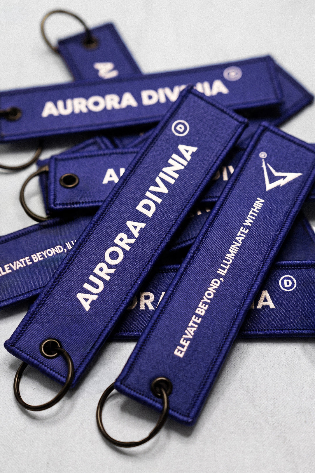 Several dark blue embroidered keychains with metal split rings, featuring white text that reads 'AURORA DIVINA' and 'ELEVATE BEYOND, ILLUMINATE WITHIN' on a light gray background.