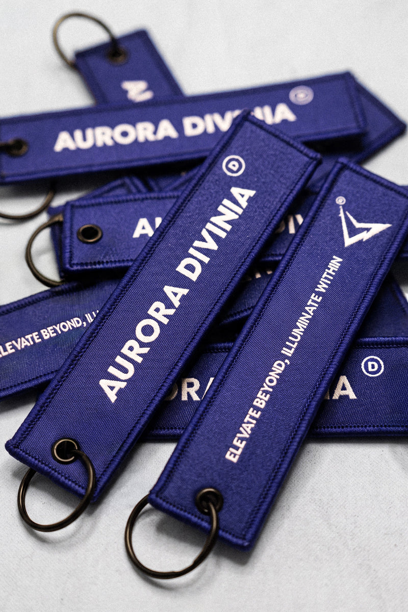 Several dark blue embroidered keychains with metal split rings, featuring white text that reads 'AURORA DIVINA' and 'ELEVATE BEYOND, ILLUMINATE WITHIN' on a light gray background.