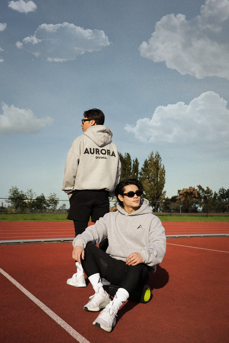 Two people wearing hoodies with 'AURORA' printed on them, sitting and standing on a red track with a grassy background.
