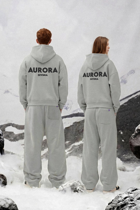 Two models from behind wearing matching light gray hoodies and sweatpants sets with 'AURORA DIVINA' printed in black on the back, set against a snowy mountain backdrop.
