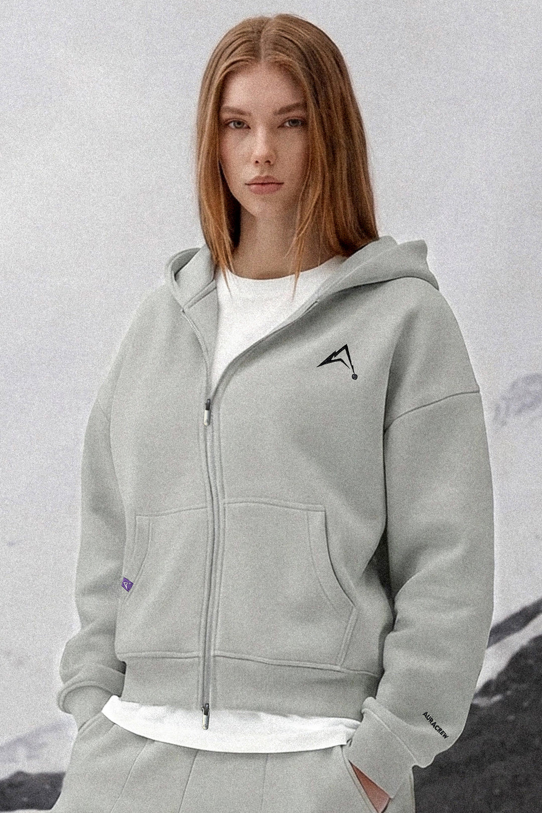 A woman wearing a light gray zip-up hoodie with a black geometric logo on the left chest, layered over a white t-shirt and matching gray sweatpants, set against a soft, light gray background.