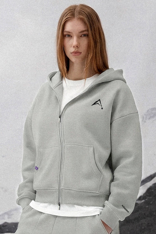 A woman wearing a light gray zip-up hoodie with a black geometric logo on the left chest, layered over a white t-shirt and matching gray sweatpants, set against a soft, light gray background.