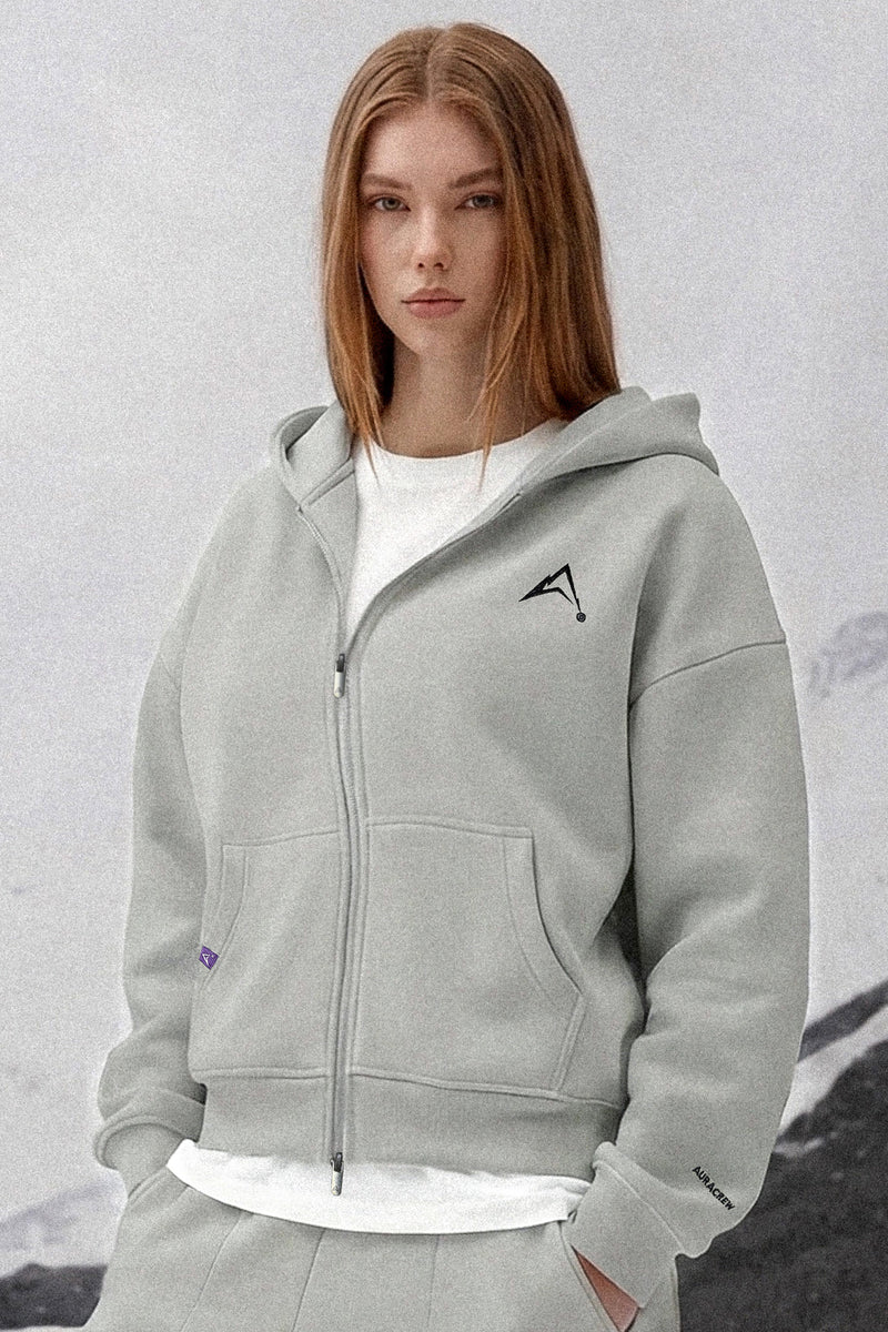 A woman wearing a light gray zip-up hoodie with a black geometric logo on the left chest, layered over a white t-shirt and matching gray sweatpants, set against a soft, light gray background.