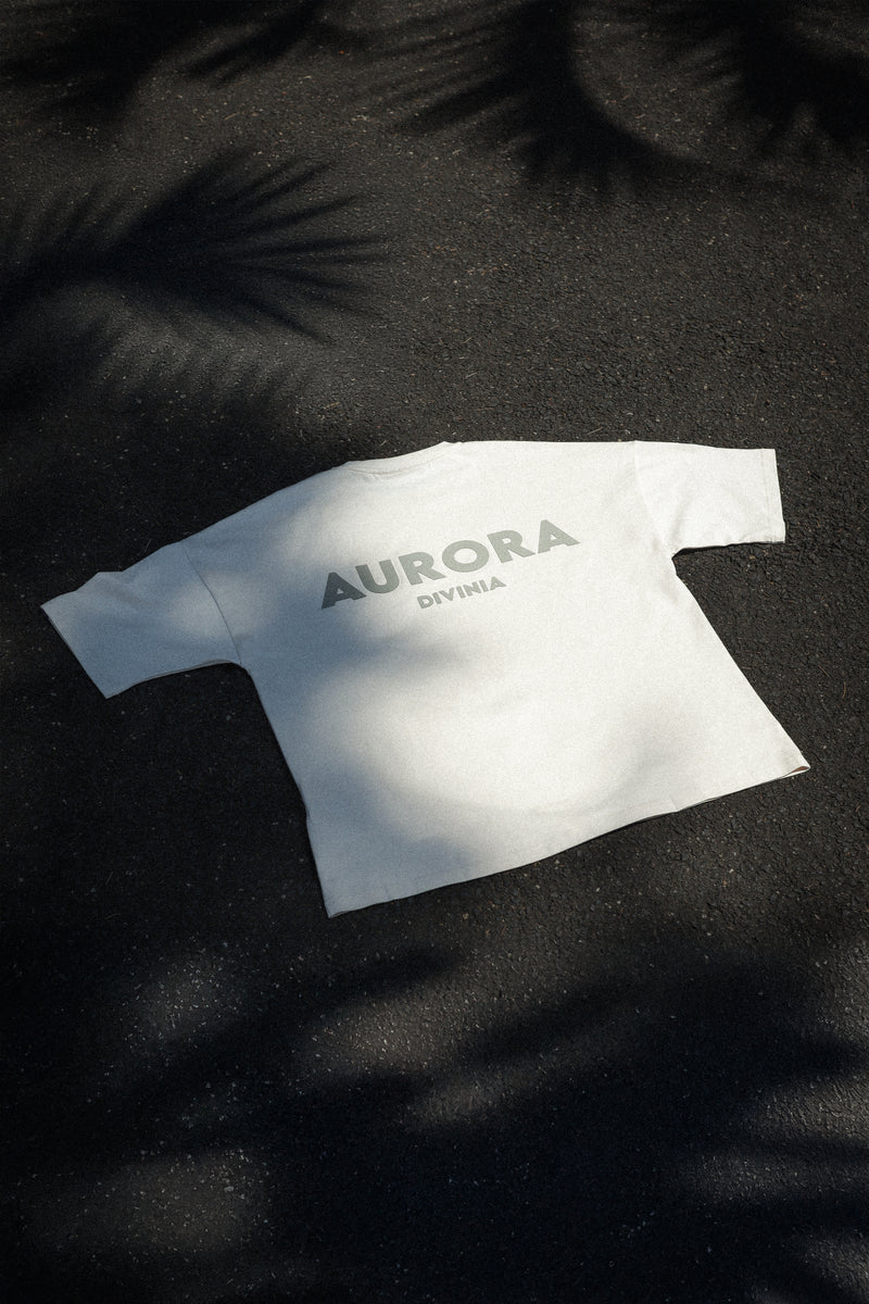 A beige crew neck t-shirt with 'AURORA Divinia' printed on the back, laid flat on a dark background with foliage around.