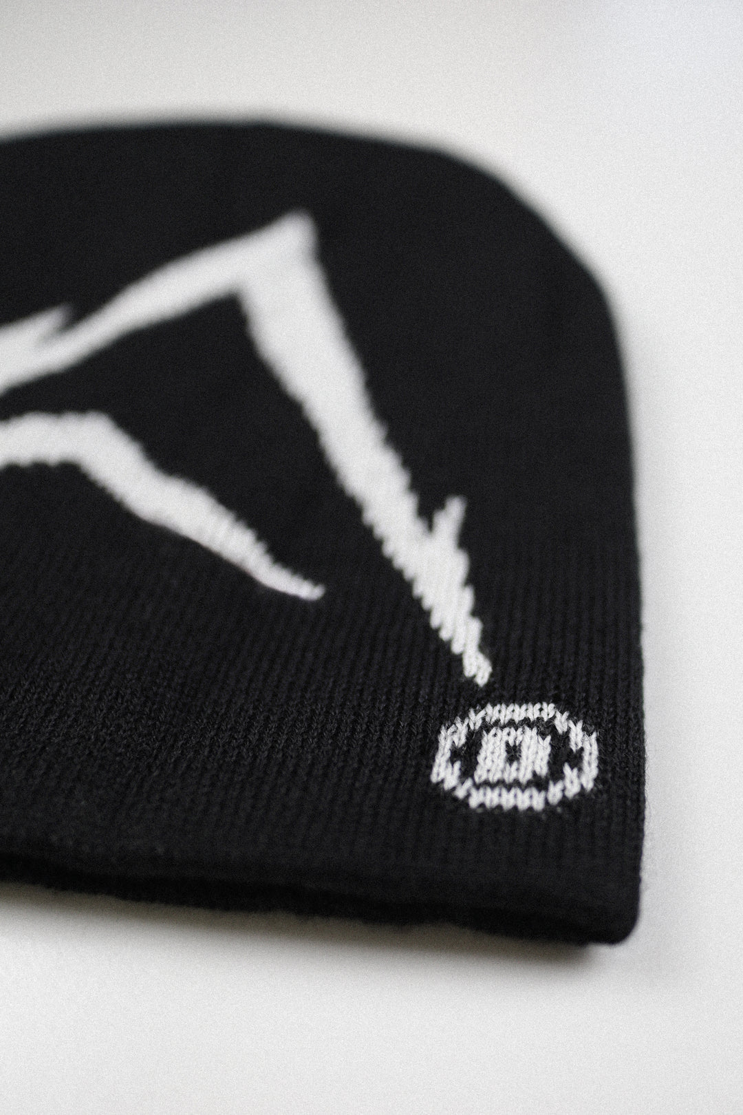 A close-up of a black knit beanie featuring a white knit mountain graphic and a small white circular logo with a stylized letter 'D' on the side, set against a plain light background.
