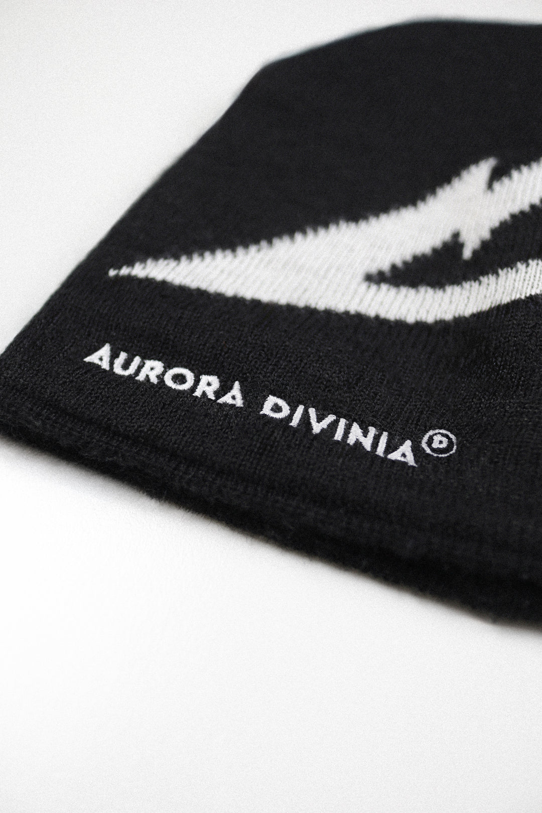 Close-up of a black knit beanie with a white embroidered lightning bolt and the text 'AURORA DIVINIA' on a white background.