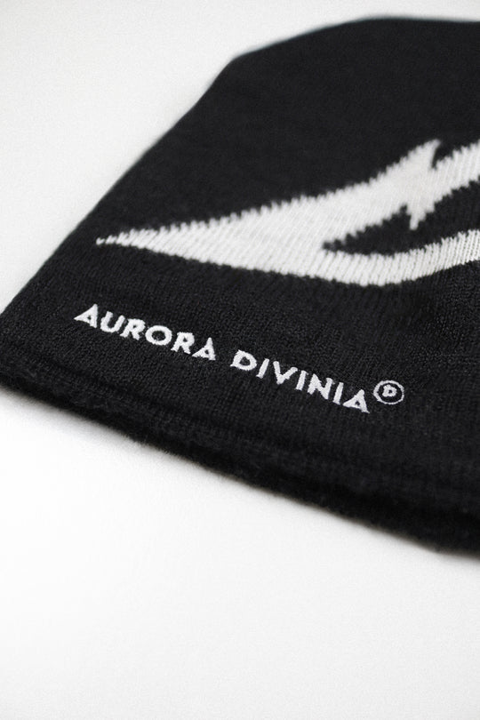 Close-up of a black knit beanie with a white embroidered lightning bolt and the text 'AURORA DIVINIA' on a white background.