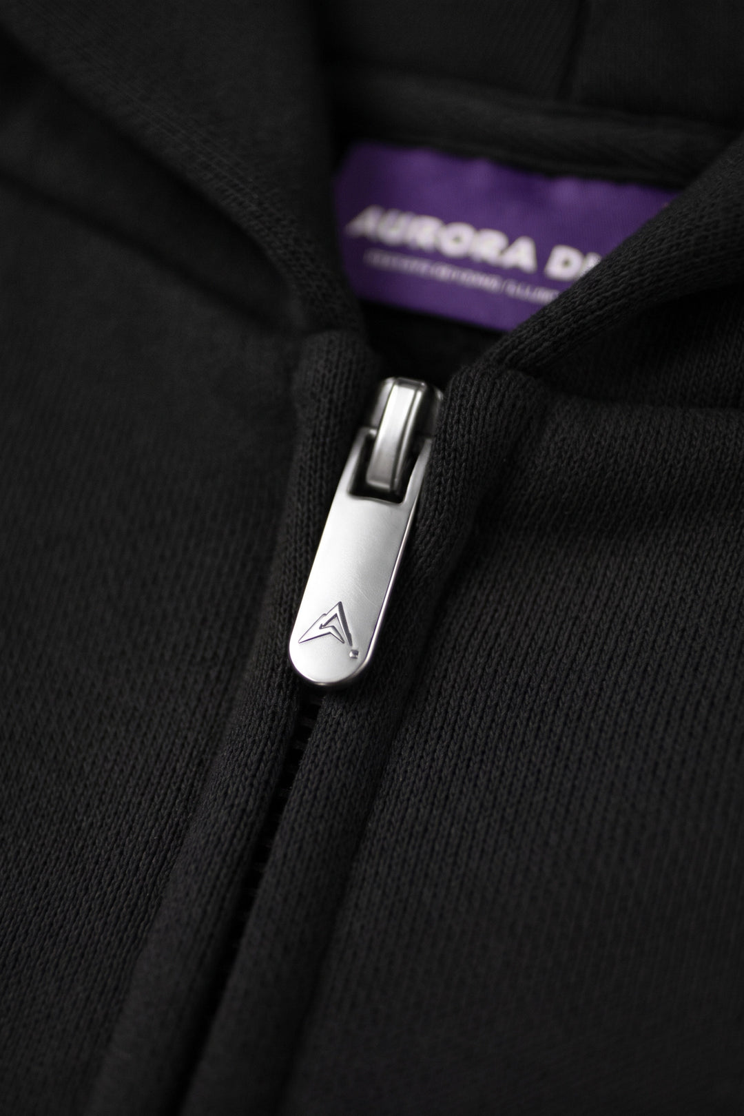 Close-up of a black zip-up hoodie with a silver metal zipper pull featuring an embossed logo, with a purple interior label reading AURORA D.