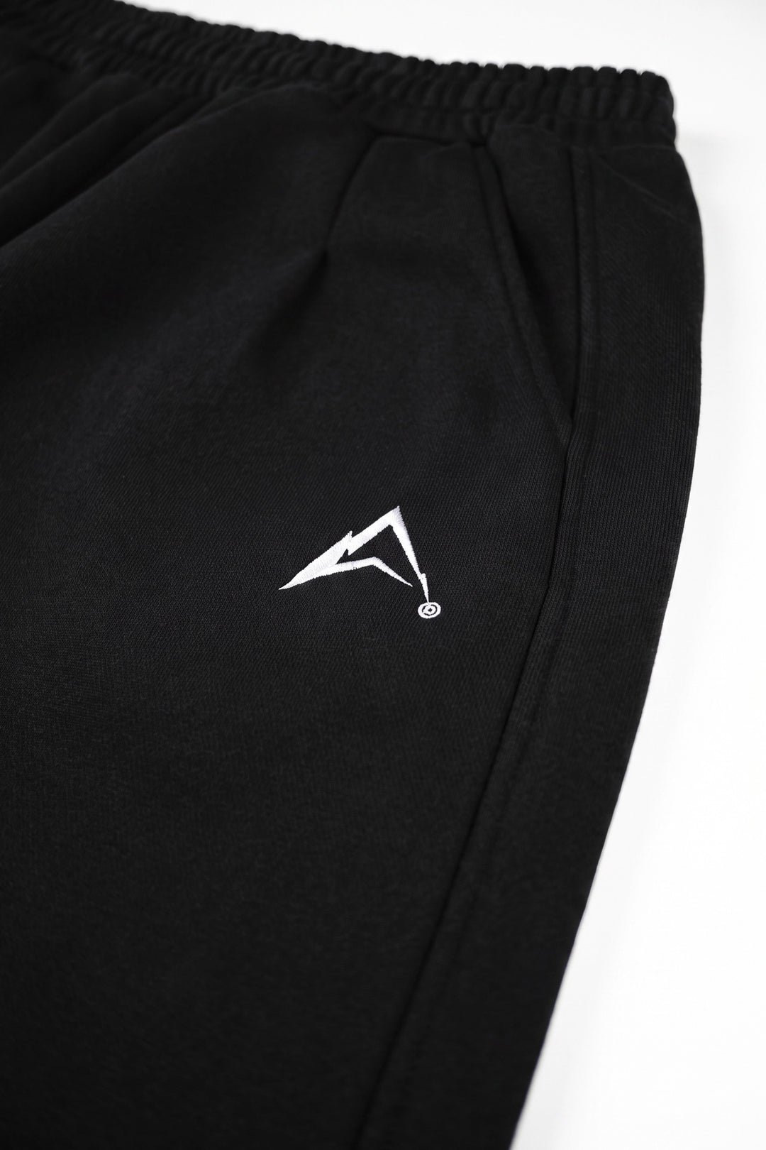 Close-up of black athletic sweatpants with an elastic waistband and a small white stylized logo on the left thigh, photographed against a white background.