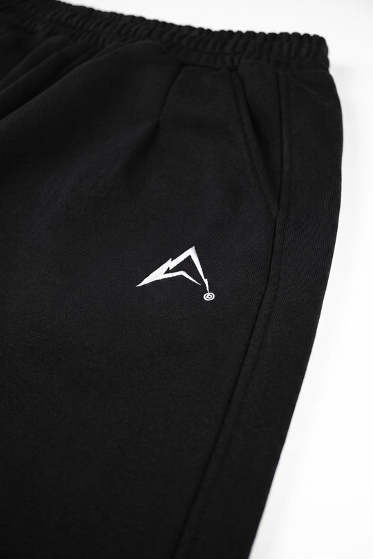 Close-up of black athletic sweatpants with an elastic waistband and a small white stylized logo on the left thigh, photographed against a white background.