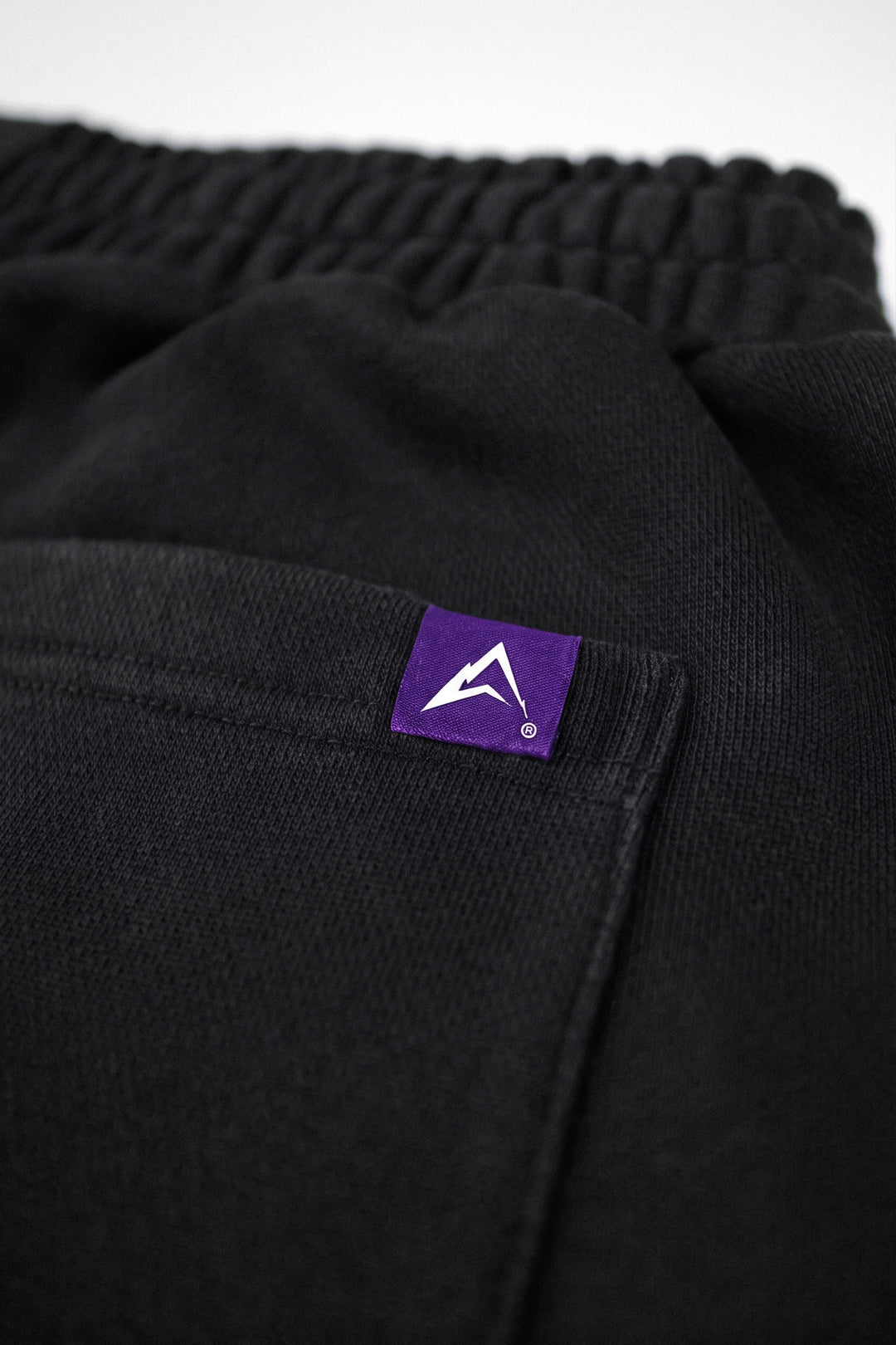 Close-up of black textured fabric with an elastic waistband and a small purple rectangular brand patch featuring a white mountain-like logo.