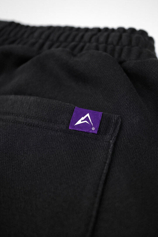 Close-up of black textured fabric with an elastic waistband and a small purple rectangular brand patch featuring a white mountain-like logo.