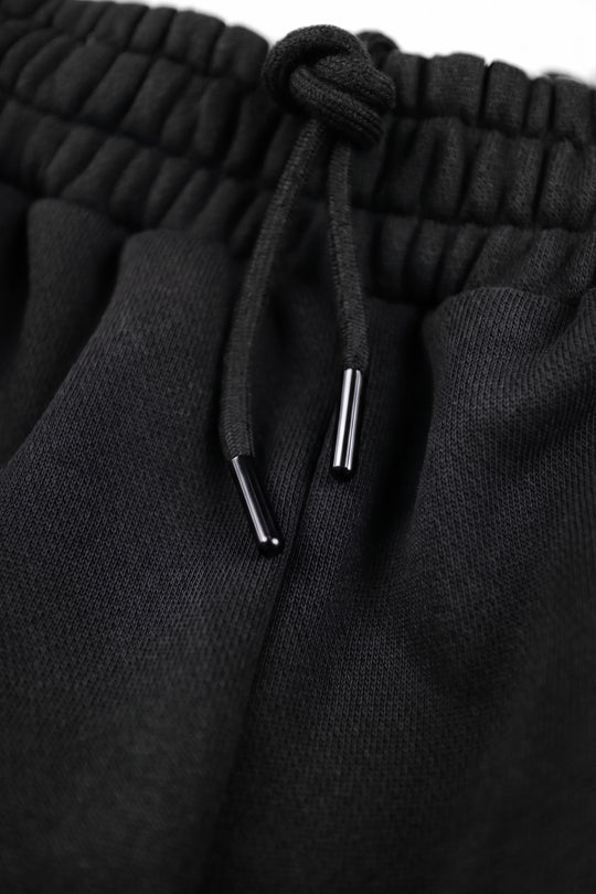 Close-up of black textured knit drawstring pants showing the elasticized waistband and black drawstring with silver metal tips.