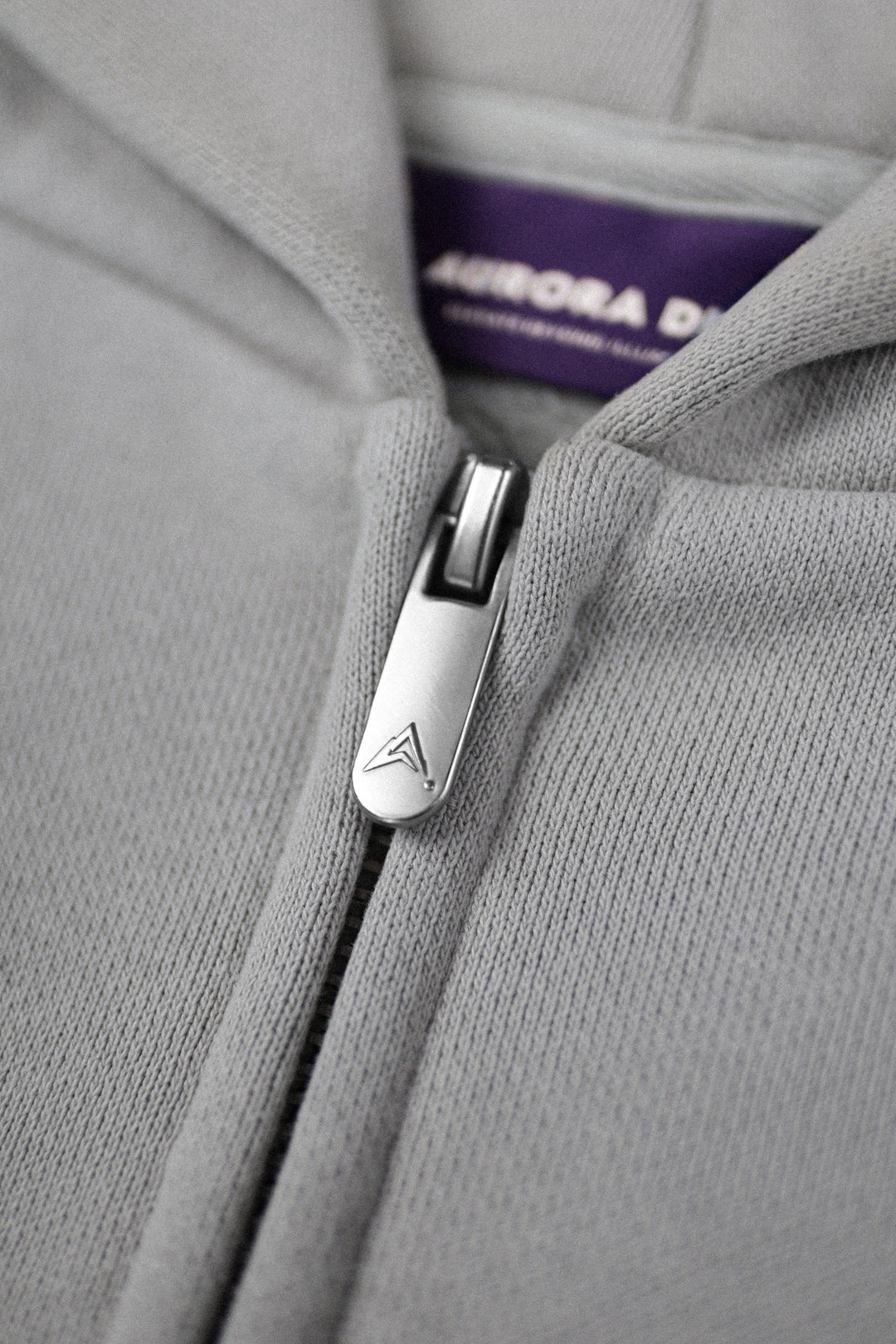 Close-up of a light gray zip-up hoodie with a silver metal zipper pull featuring a stylized 'A' logo, and a purple interior neck label reading AURORA DIVINIA.