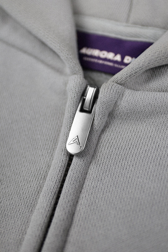 Close-up of a light gray zip-up hoodie with a silver metal zipper pull featuring a stylized 'A' logo, and a purple interior neck label reading AURORA DIVINIA.