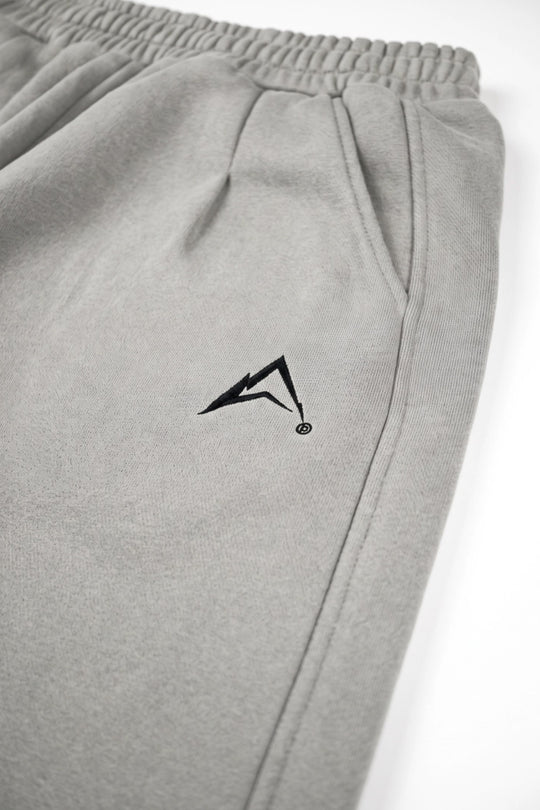 Close-up of light gray sweatpants with an elastic waistband and a black geometric logo on the left thigh, photographed against a white background.