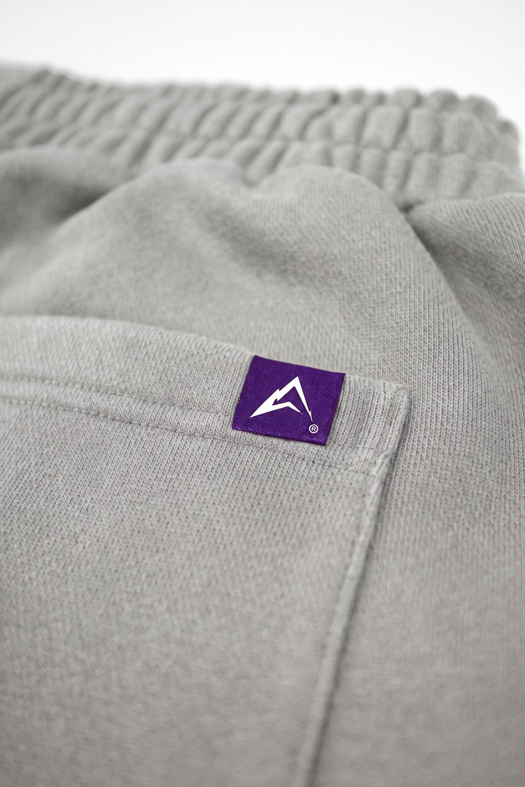 Close-up of light gray heather sweatpants showing a side pocket with a small purple square brand tag featuring a white mountain logo.