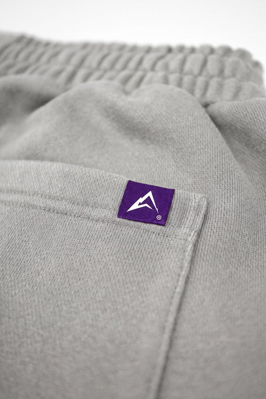 Close-up of light gray heather sweatpants showing a side pocket with a small purple square brand tag featuring a white mountain logo.
