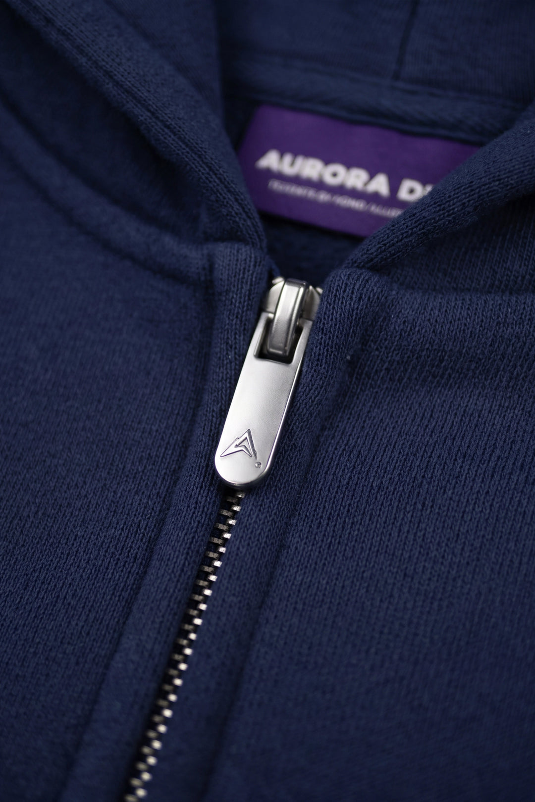 Close-up of a navy blue zip-up hoodie with a silver metal zipper pull and a purple interior neck label reading AURORA DESIGN.