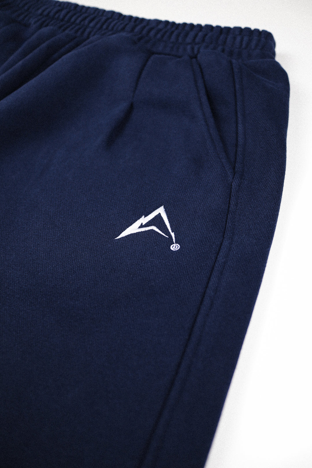 Close-up of navy blue sweatpants with a white stylized logo on the thigh, shown against a plain white background.