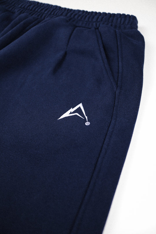 Close-up of navy blue sweatpants with a white stylized logo on the thigh, shown against a plain white background.