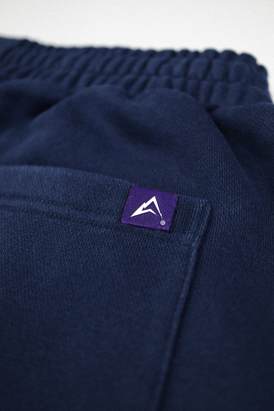 Close-up of dark navy knit fabric with an elastic waistband and a small purple brand patch featuring a white mountain logo on the side pocket.