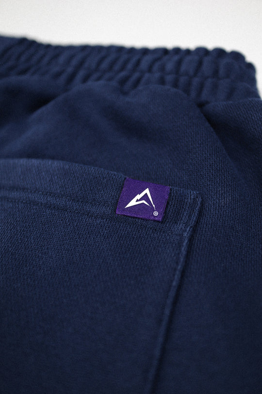 Close-up of dark navy knit fabric with an elastic waistband and a small purple brand patch featuring a white mountain logo on the side pocket.